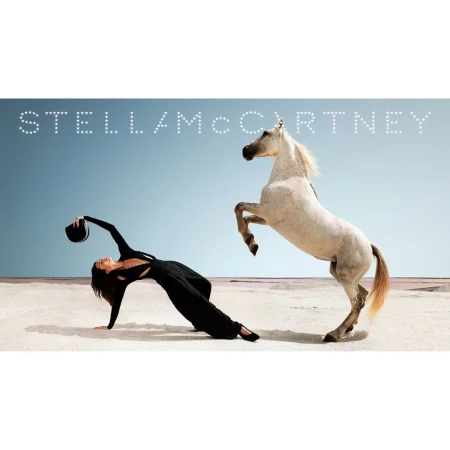 A woman in black clothing arches backward on a light sandy beach next to a rearing white horse beneath a pale sky.