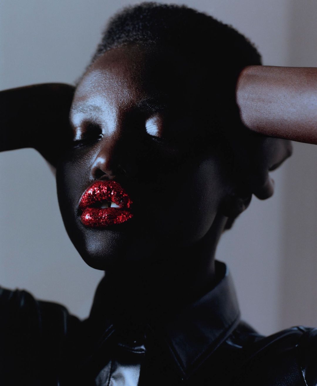 Close-up portrait of a woman with dark skin and red glitter lipstick, holding her hands behind her head, against a neutral background.