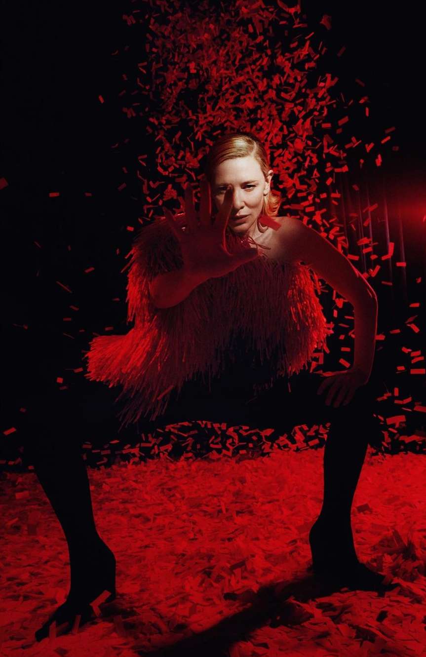 Actress Cate Blanchett poses in a studio with a white fringe top and confetti-like pieces against a dark red background.