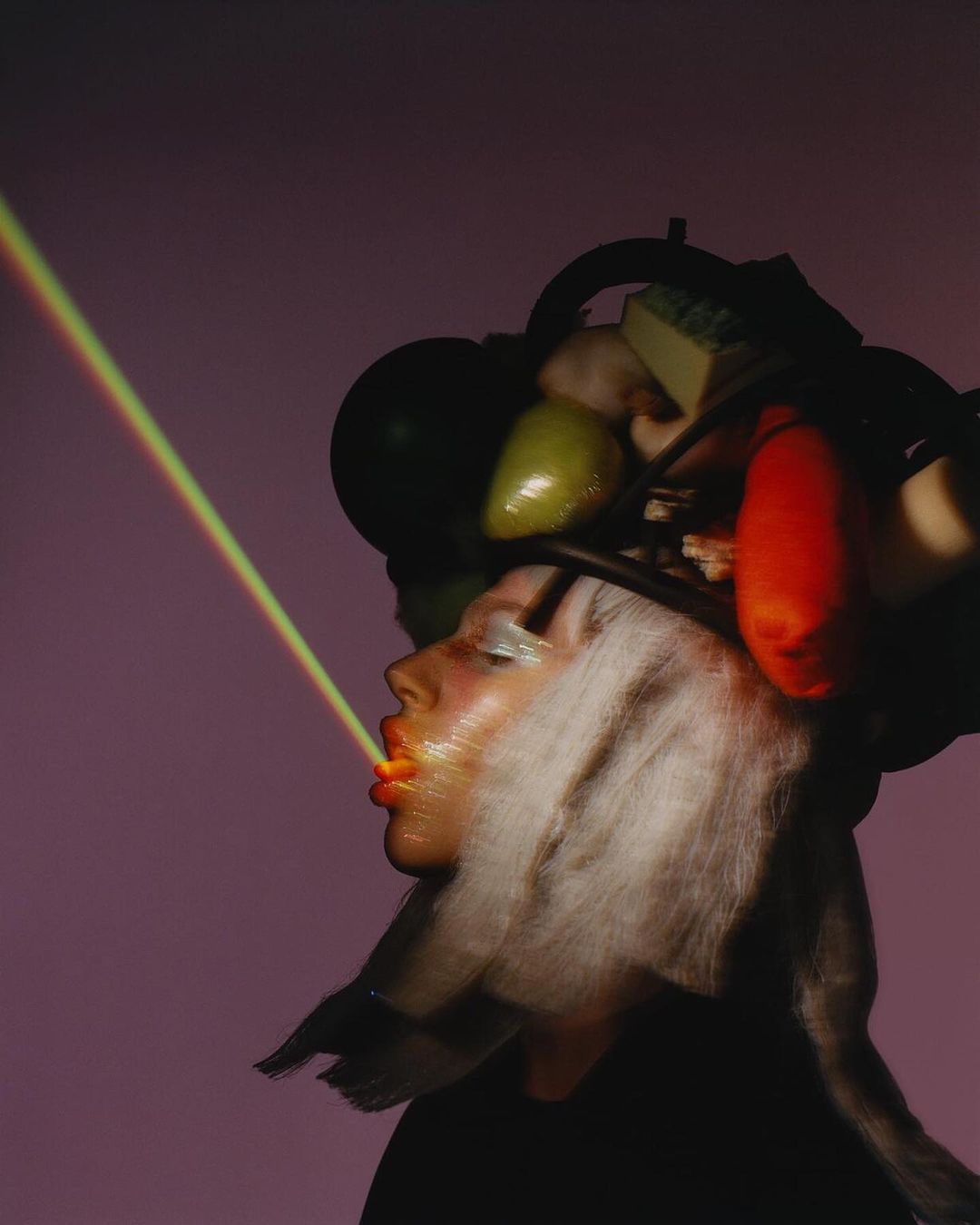 Profile shot of a person with fruits and objects on their head, blowing a laser beam from their mouth against a purple background.