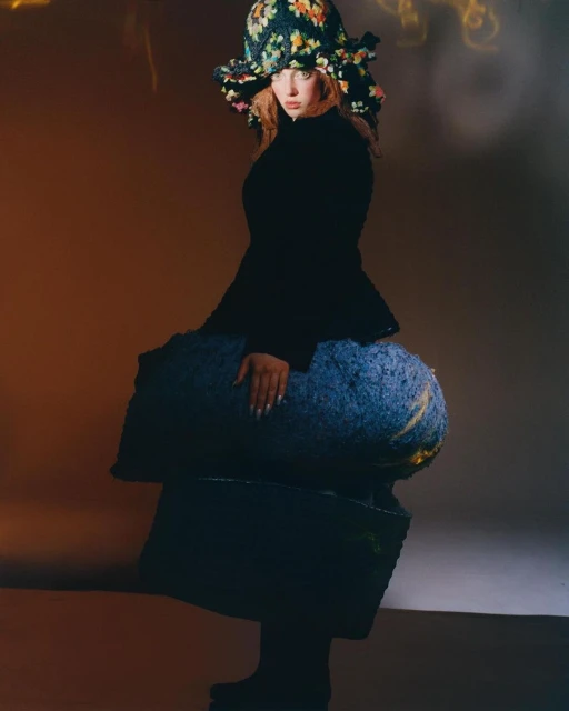 A woman wearing a crocheted hat and black jacket poses in a studio, sitting on an abstract blue and yellow platform.