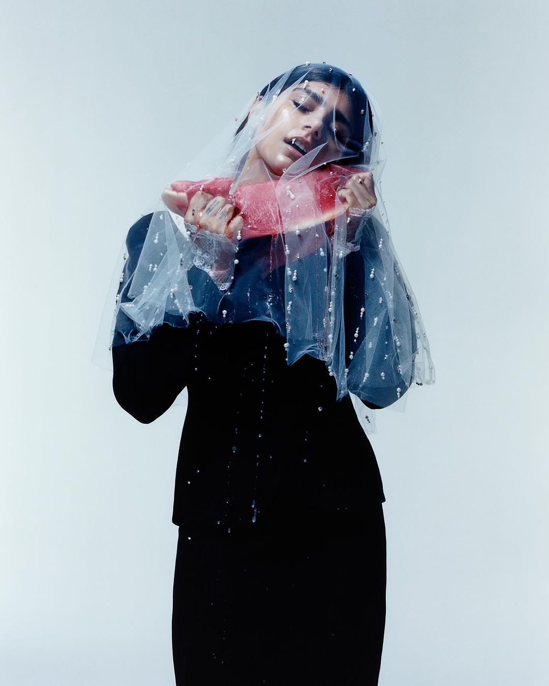 Portrait of a woman in a black dress and veil, holding a piece of watermelon with water droplets on a light grey background.