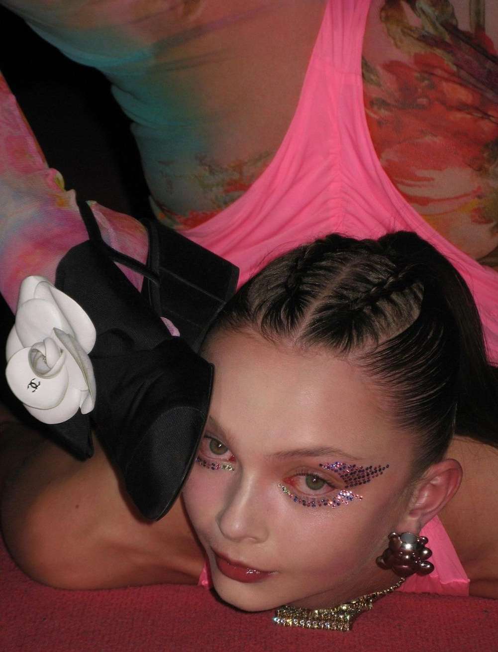 A young woman poses with black high heel shoes adorned with a white flower detail, makeup, and jewelry against a colorful background.