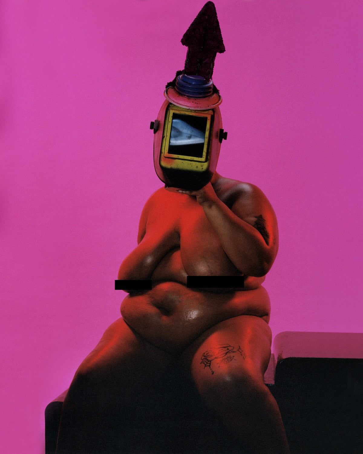 Adult nude figure sitting with welding mask and arrow headpiece against a pink background in a studio setting. The figure has a tattoo.