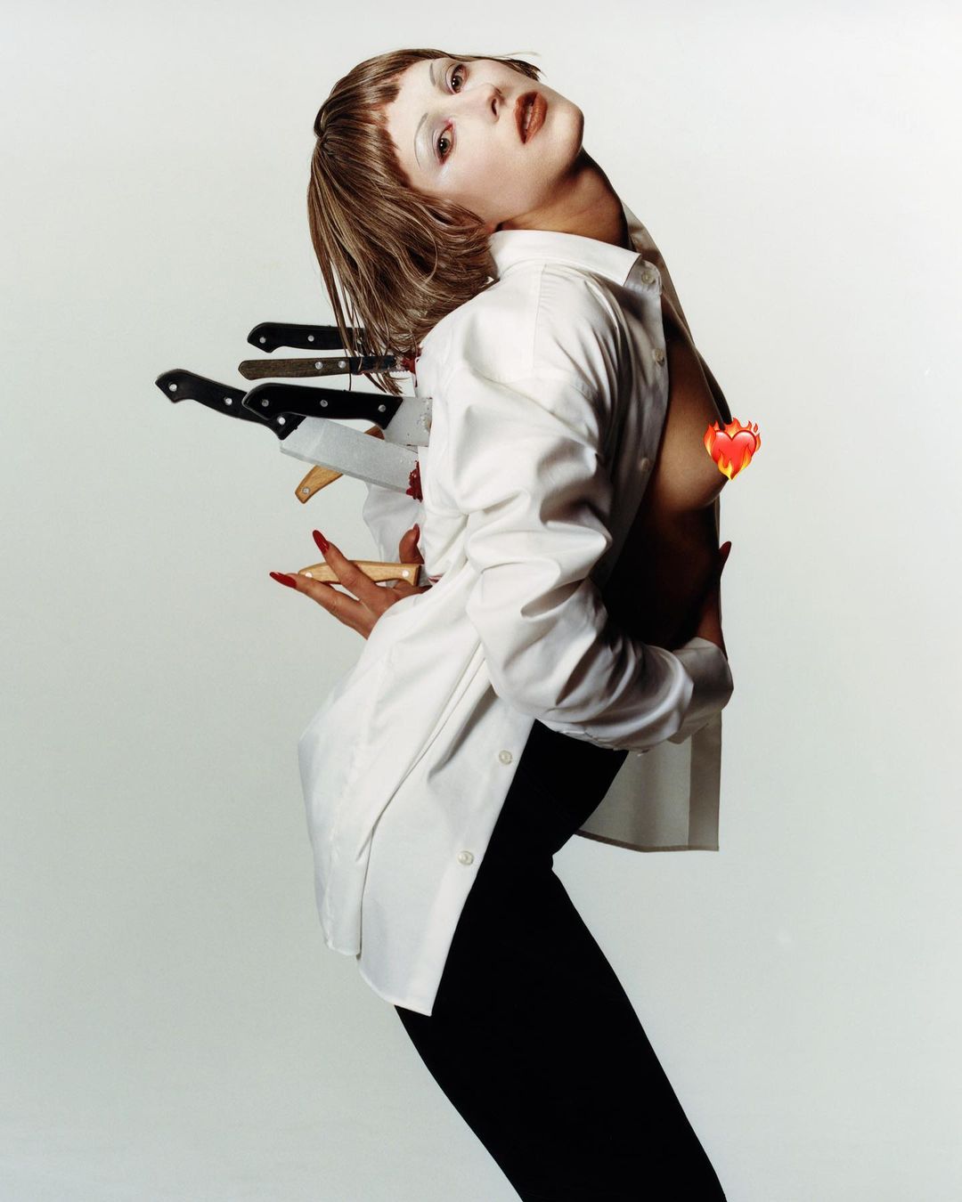 Dramatic studio shot of a woman with fair skin wearing a white button-down shirt and black pants, posing with knives around her body.