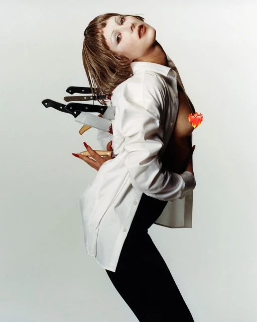 Dramatic studio shot of a woman with fair skin wearing a white button-down shirt and black pants, posing with knives around her body.