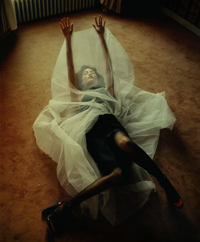 Woman lying on a brown carpet under sheer white tulle fabric with hands raised, photographed by Harley Weir