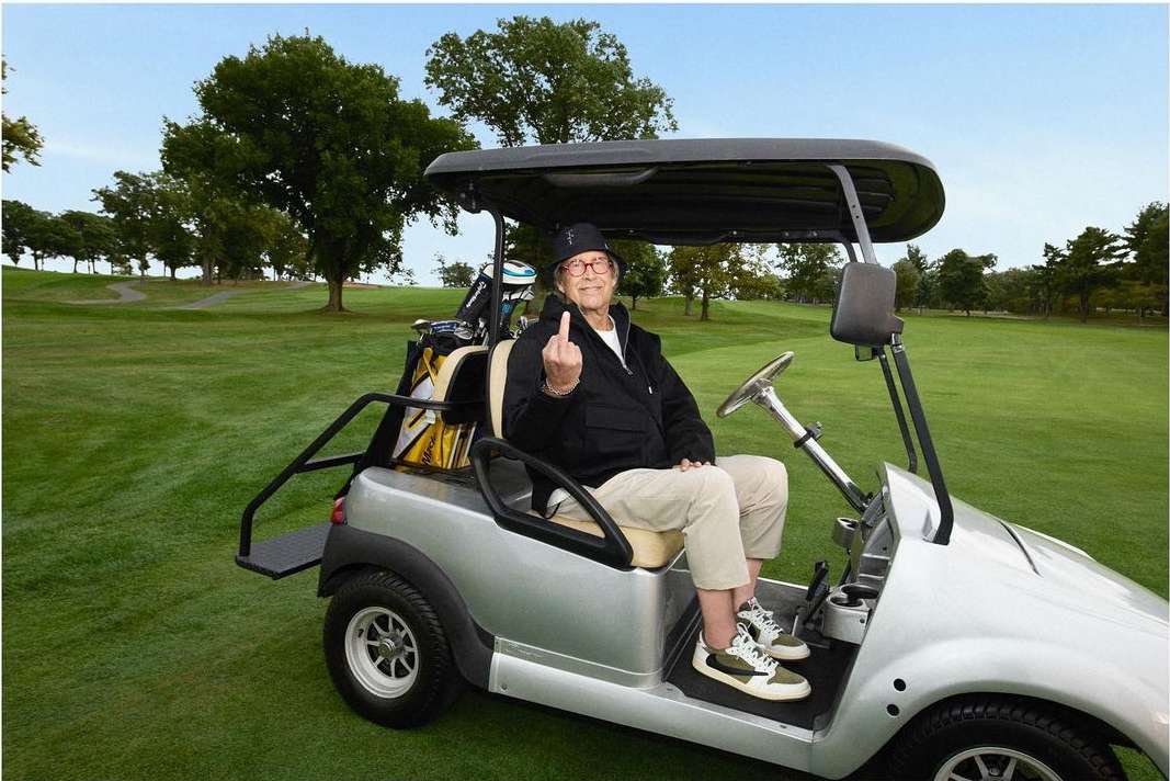 An elderly man sits in a golf cart on a lush green golf course, giving the middle finger with a smile and wearing glasses.