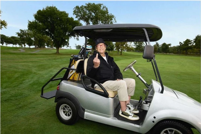 An elderly man sits in a golf cart on a lush green golf course, giving the middle finger with a smile and wearing glasses.