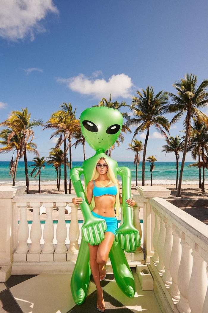 A blonde woman in a blue bikini poses with a large, green, inflatable alien on a balcony overlooking a beach with palm trees and blue sky.