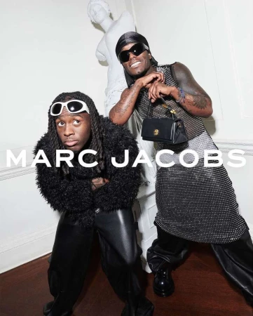 Two men posing together in stylish apparel with sunglasses, next to a classical marble statue against a white wall.