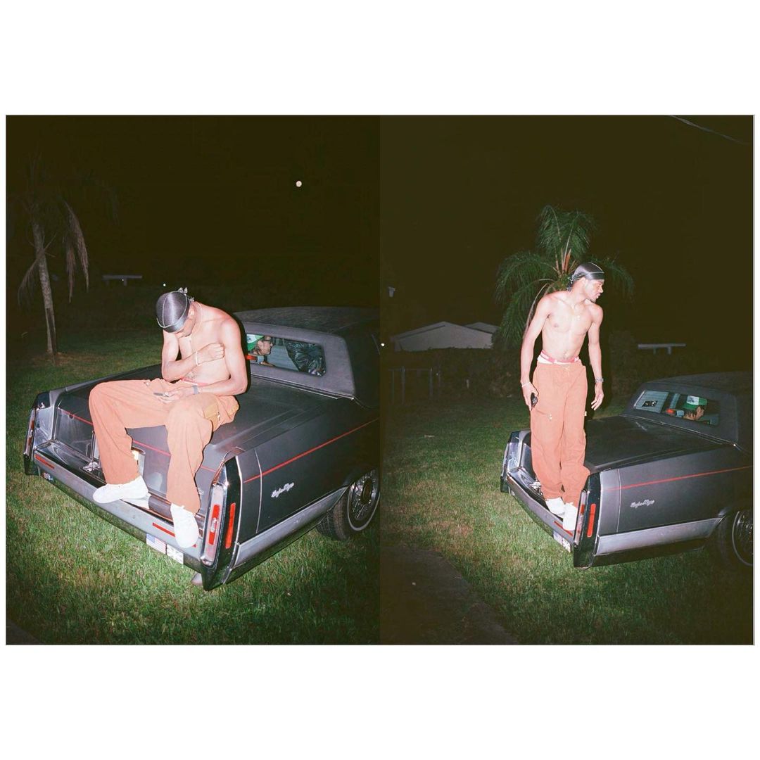 A young man in an orange outfit poses on the trunk of a vintage car at night, near palm trees and a building.