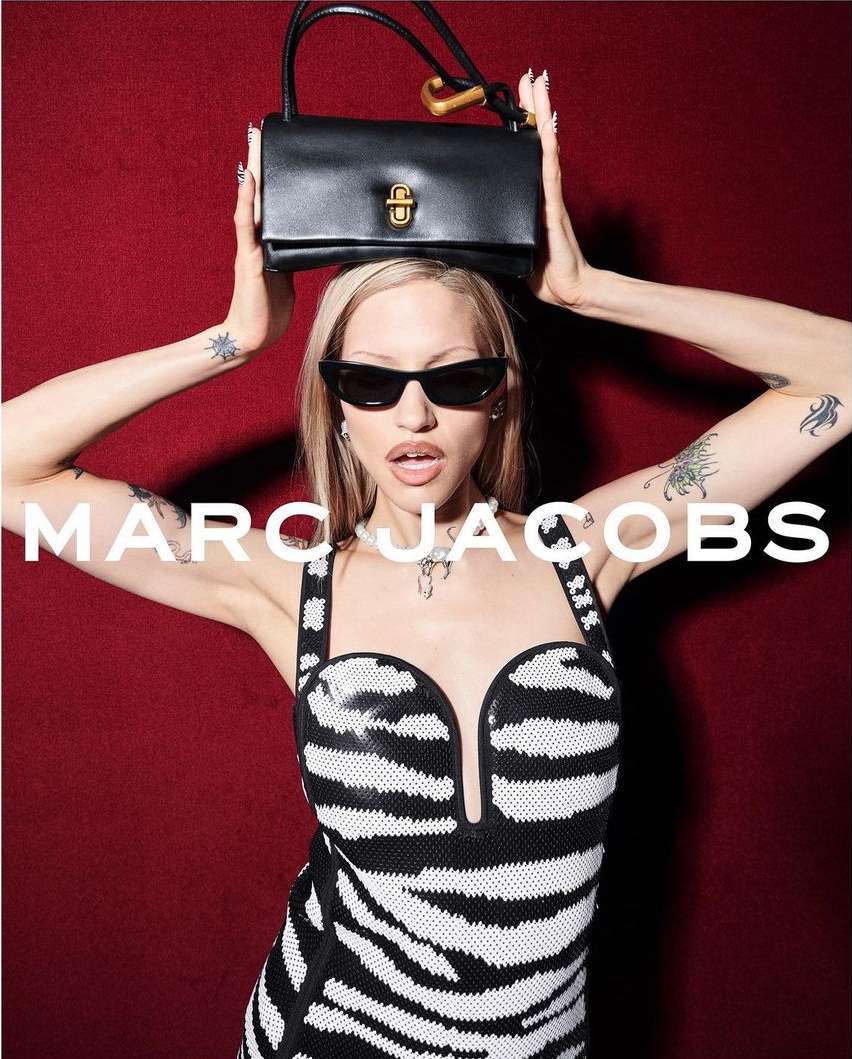 A model poses in a zebra-striped dress with a black leather bag held over her head against a red background, showcasing a Marc Jacobs product.