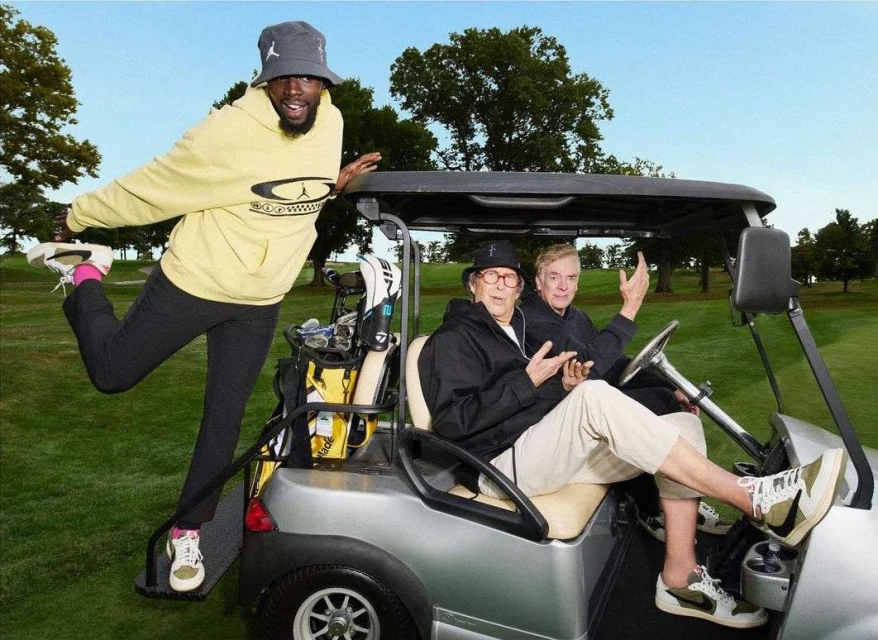 Two older men ride in a golf cart while a younger man stands on the back footrest, on a green golf course with trees and blue sky.