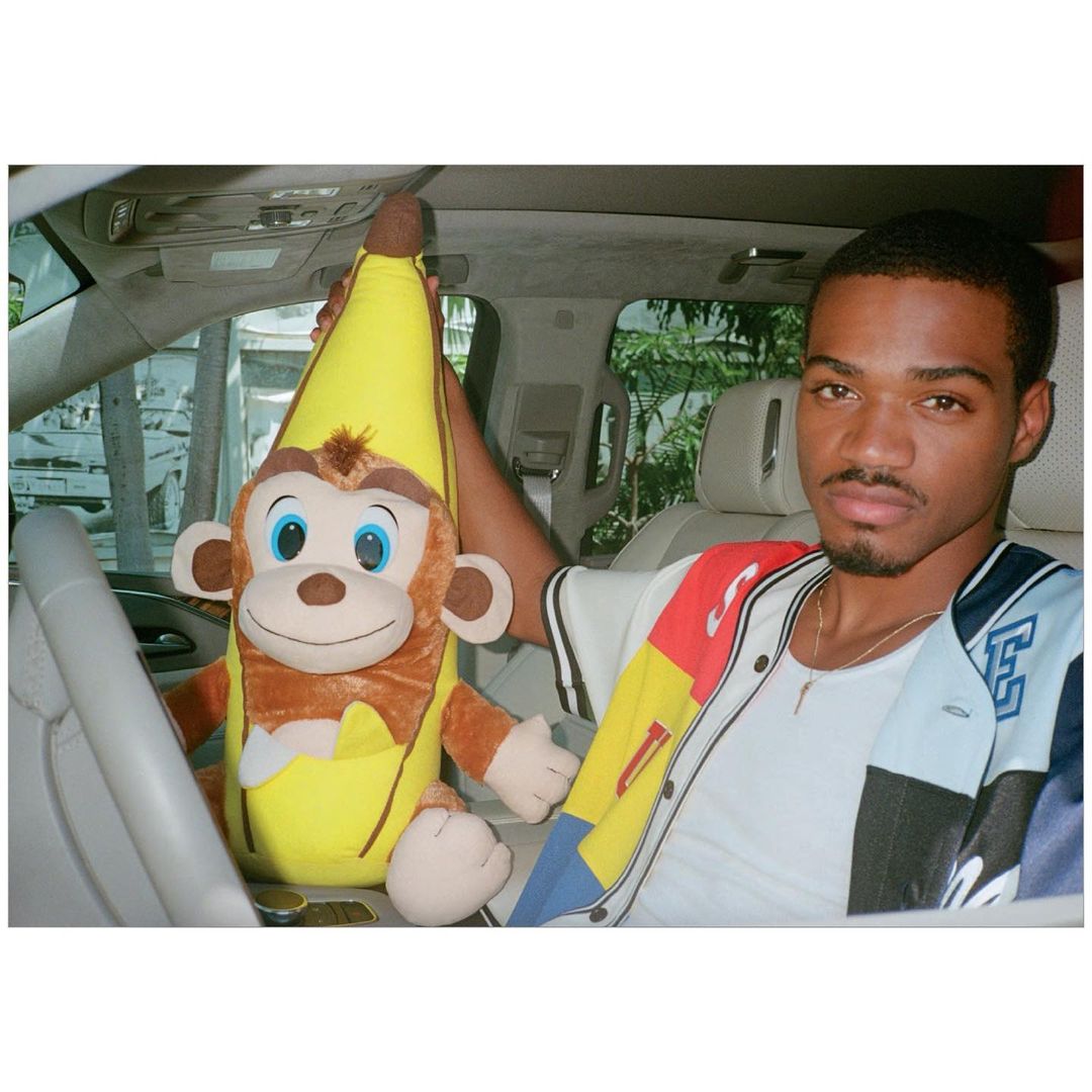 A young man sitting in a car holding a monkey-banana plush toy, wearing a baseball jacket with a white car interior visible.