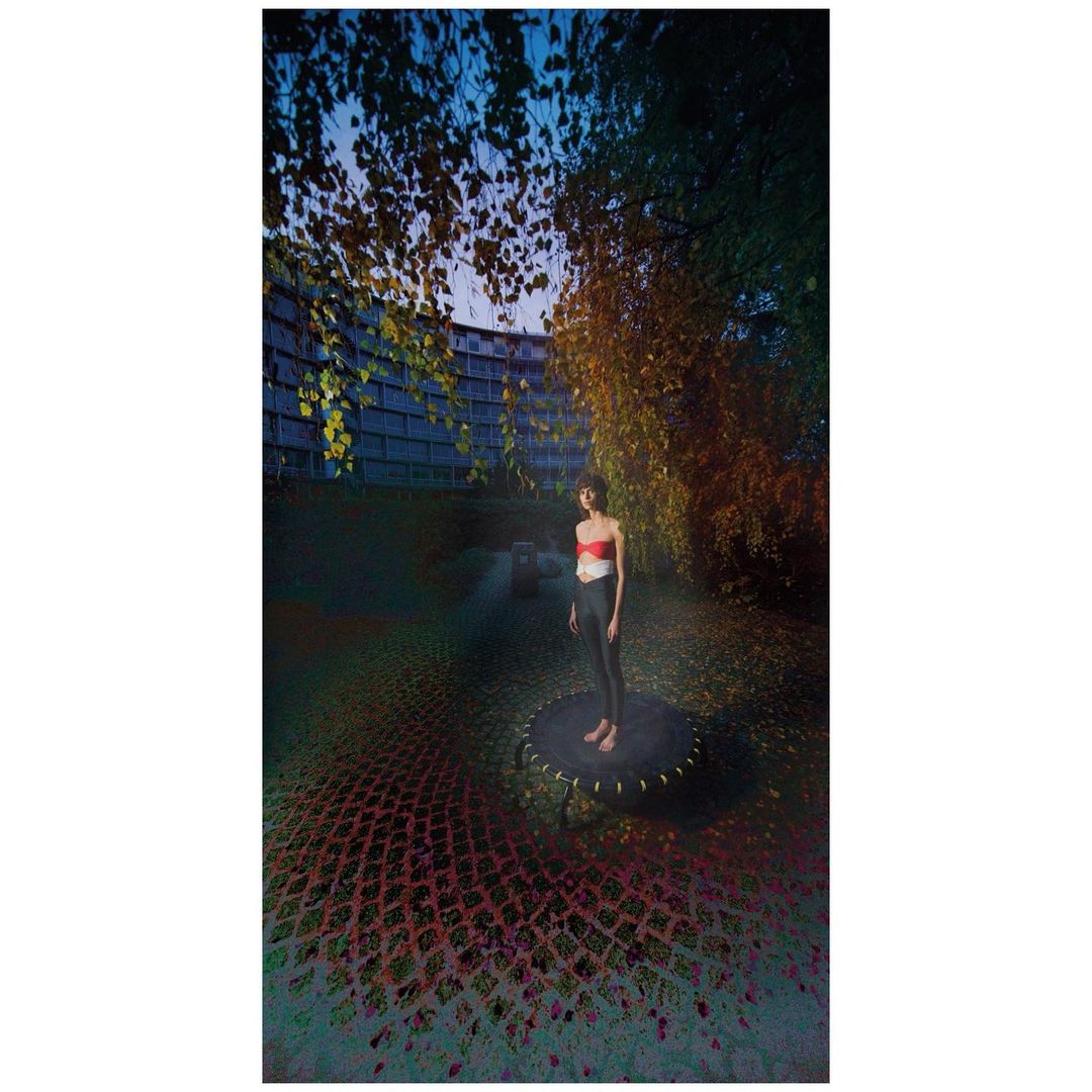 Woman in activewear stands on trampoline outdoors at dusk; architecture and foliage fill the background in a surreal fashion image.