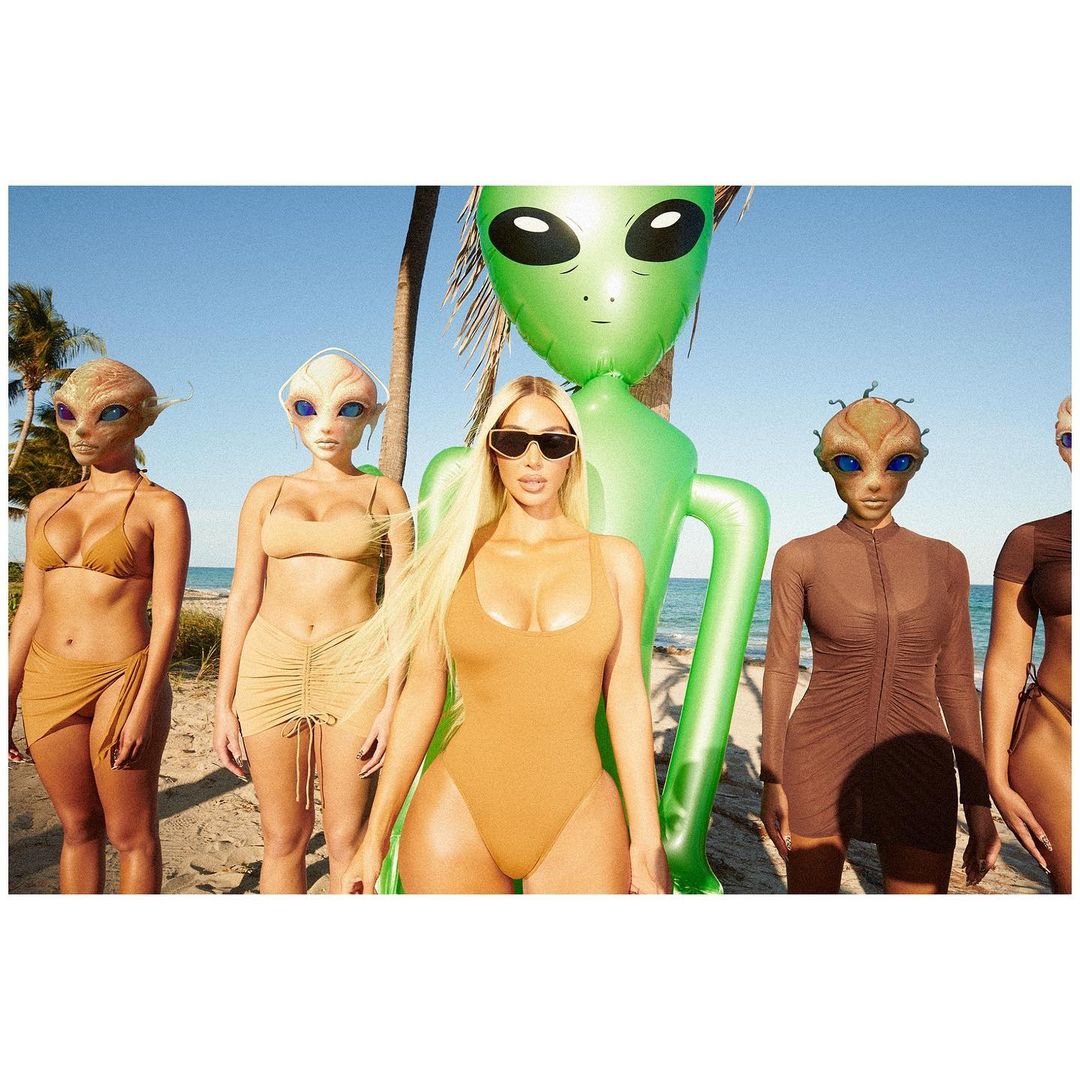 Kim Kardashian posing with women in a tan swimwear on a beach. The women appear to have alien heads, while an inflatable alien stands nearby.