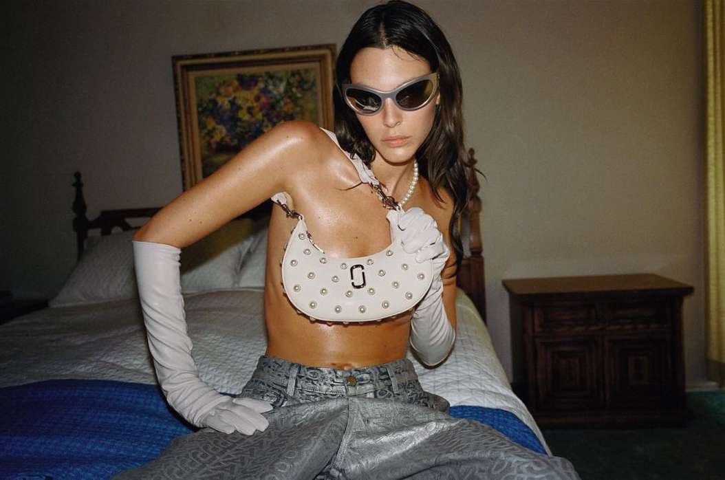 Fashion shot featuring a woman in a silver outfit and sunglasses posing with a white handbag on a bed in an indoor setting.