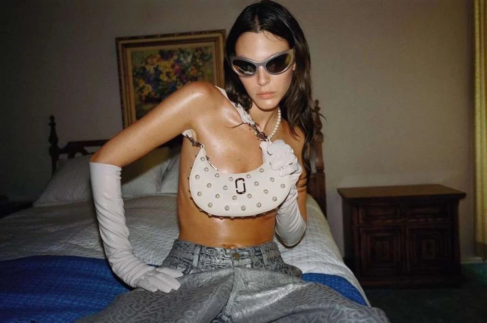 Fashion shot featuring a woman in a silver outfit and sunglasses posing with a white handbag on a bed in an indoor setting.