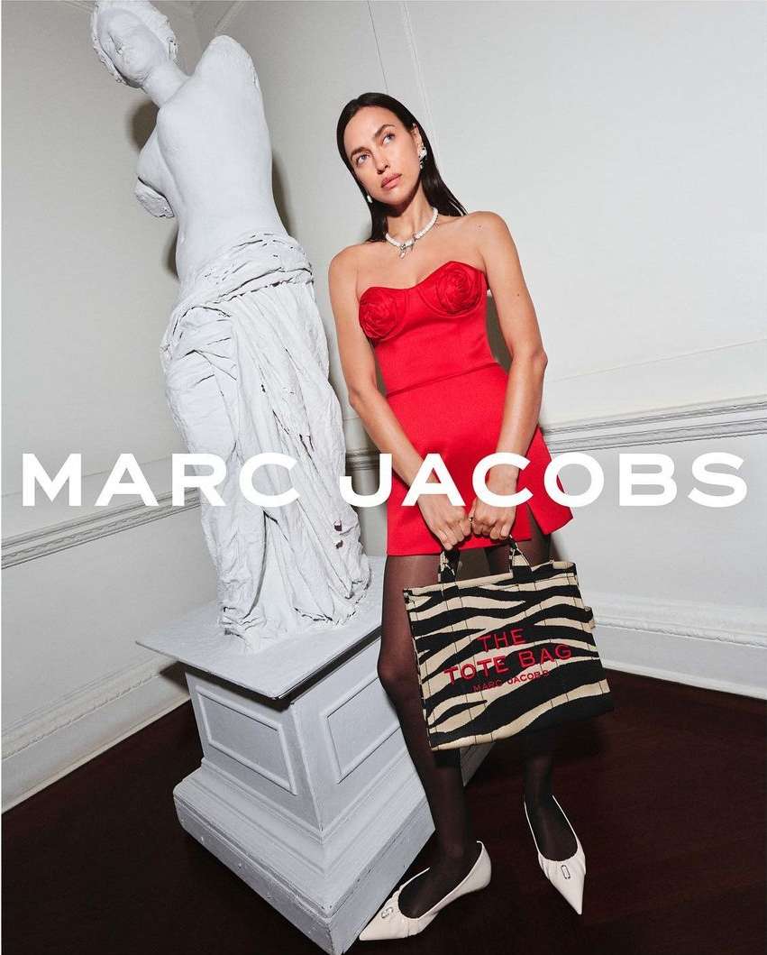 Elegant woman in red dress posing with a Marc Jacobs tote bag next to a white statue indoors, showcasing a fashion-forward aesthetic.