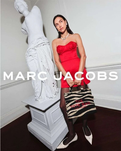 Elegant woman in red dress posing with a Marc Jacobs tote bag next to a white statue indoors, showcasing a fashion-forward aesthetic.