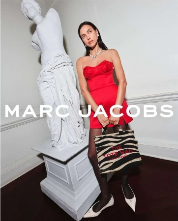 Elegant woman in red dress posing with a Marc Jacobs tote bag next to a white statue indoors, showcasing a fashion-forward aesthetic.