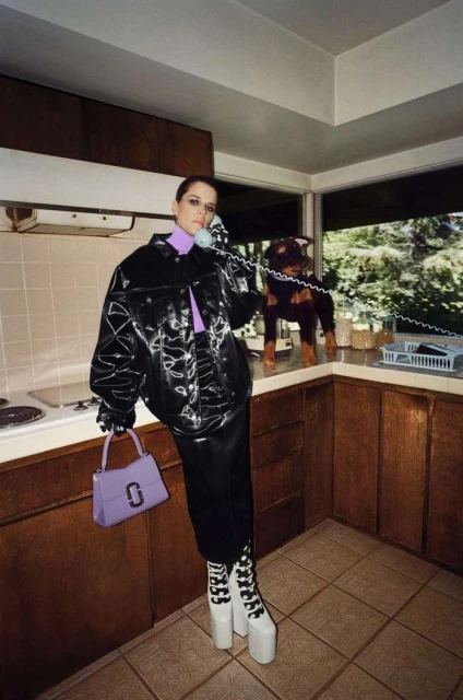 Woman in vinyl jacket, skirt, and platform boots talking on the phone indoors, with a dog on the kitchen counter and holding purse.