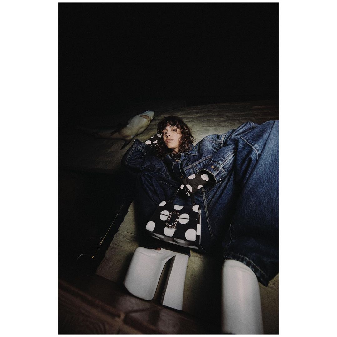 Fashion photo of a woman in a denim jacket and polka dot gloves holding a polka dot purse, sitting atop white platform boots.