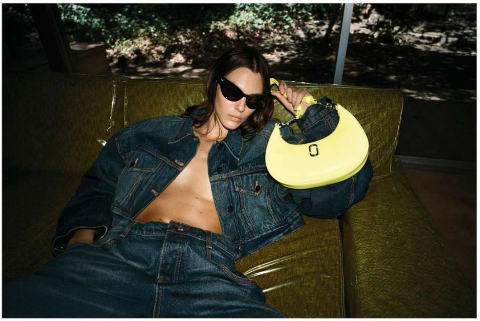 A woman in a denim outfit reclines on a sofa with a yellow purse, captured in an indoor setting with natural lighting.