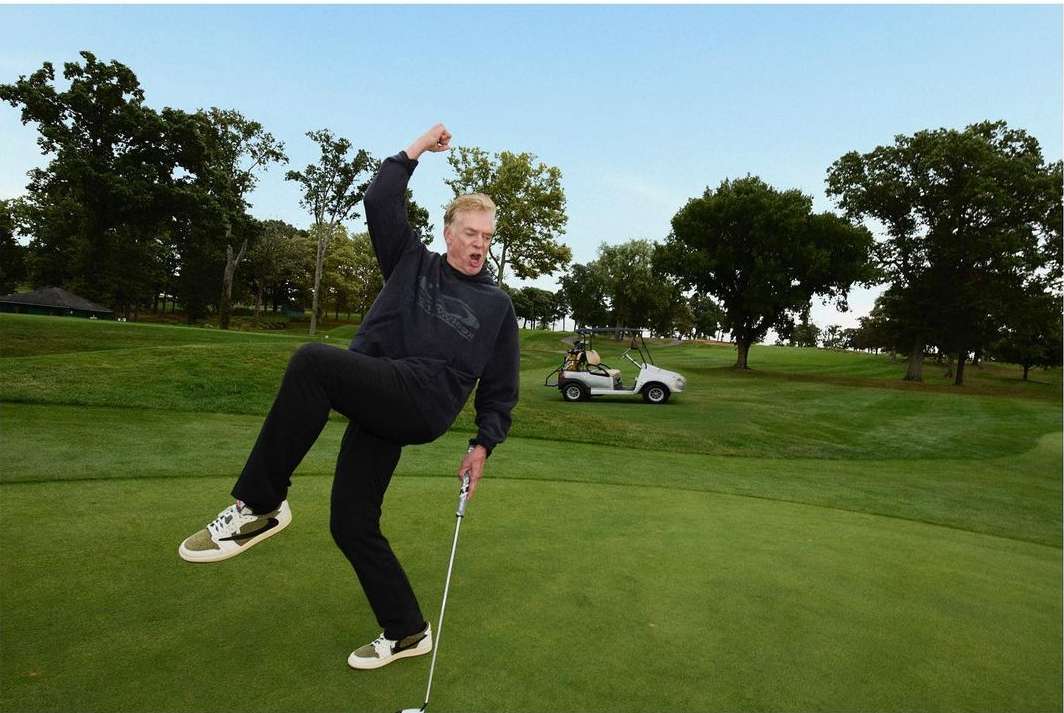 An adult male golfer in mid-swing on a green golf course on a sunny day, wearing black pants and top, with trees and a golf cart in the background.