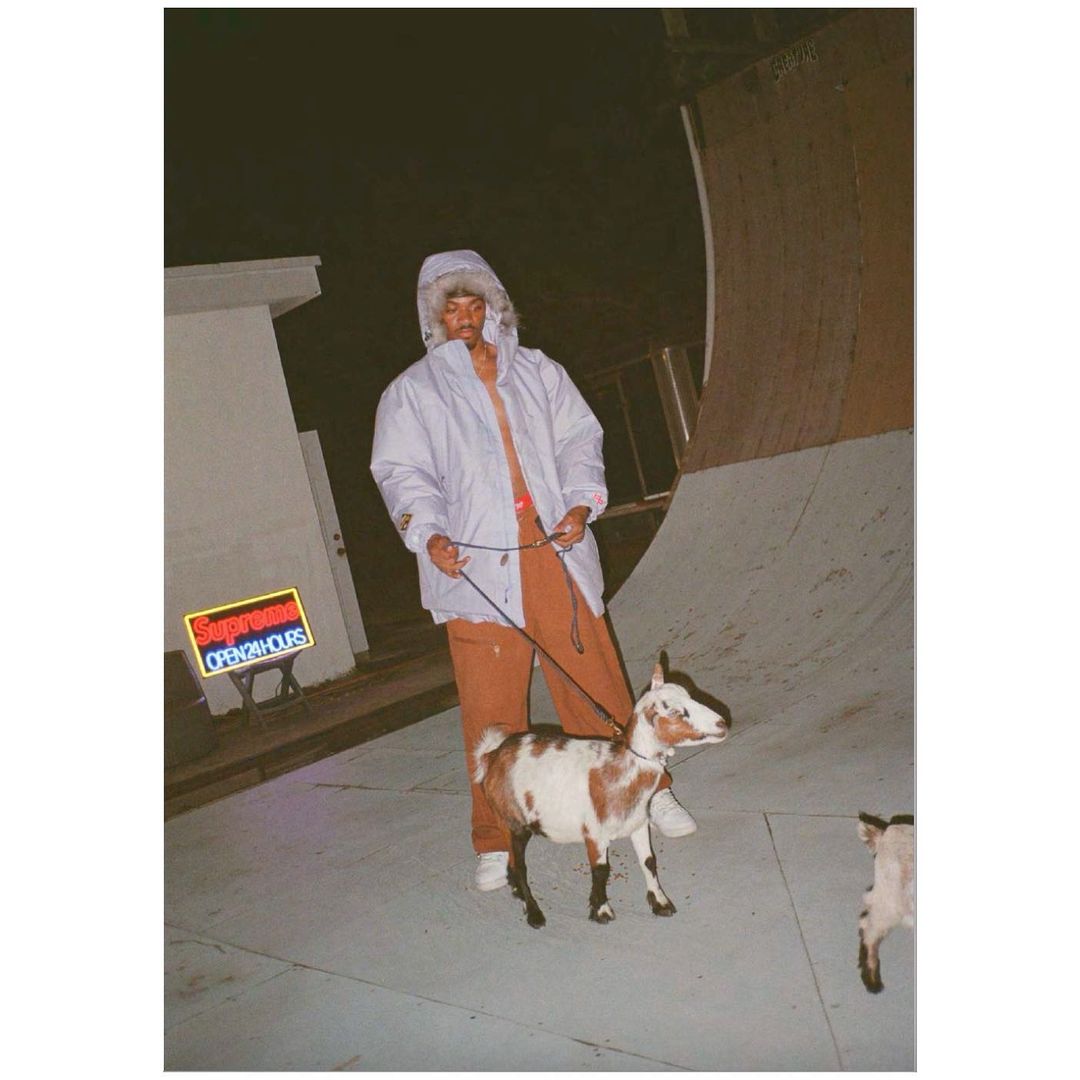 A man wearing a hooded jacket is walking a goat on a leash on a concrete surface next to a skate ramp at night, with a neon sign.
