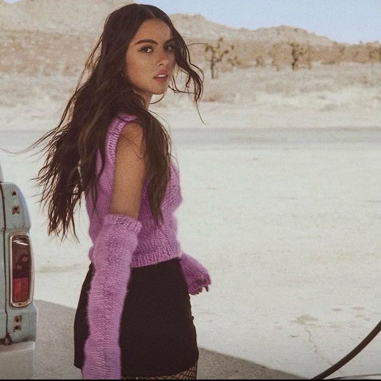 Young woman in lilac knitted top and arm warmers poses in desert landscape, next to a vintage truck, looking over her shoulder.