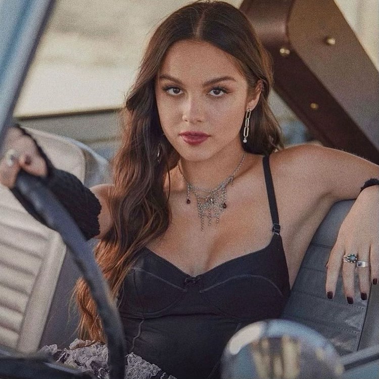 Young woman with long wavy brown hair, wearing a black top and jewelry, driving an old car. She has a serious expression.