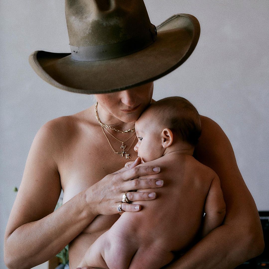 Woman wearing hat and jewelry holds a baby in an indoor setting against a plain wall, with a shallow depth of field.