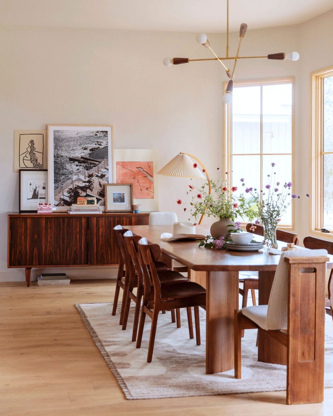 Dining room interior with wooden dining table set with flowers, dark wood sideboard, and modern lighting in a bright, neutral space.