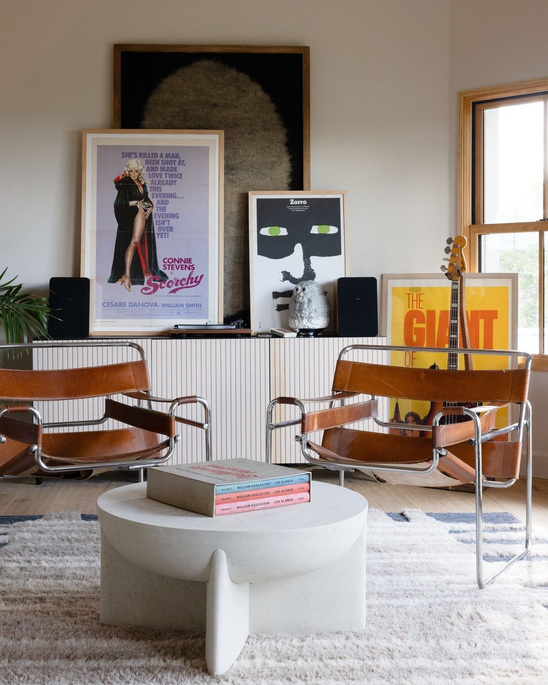 Living room interior with a round white coffee table with books, modern chairs, posters, guitar, and speakers against a white wall.