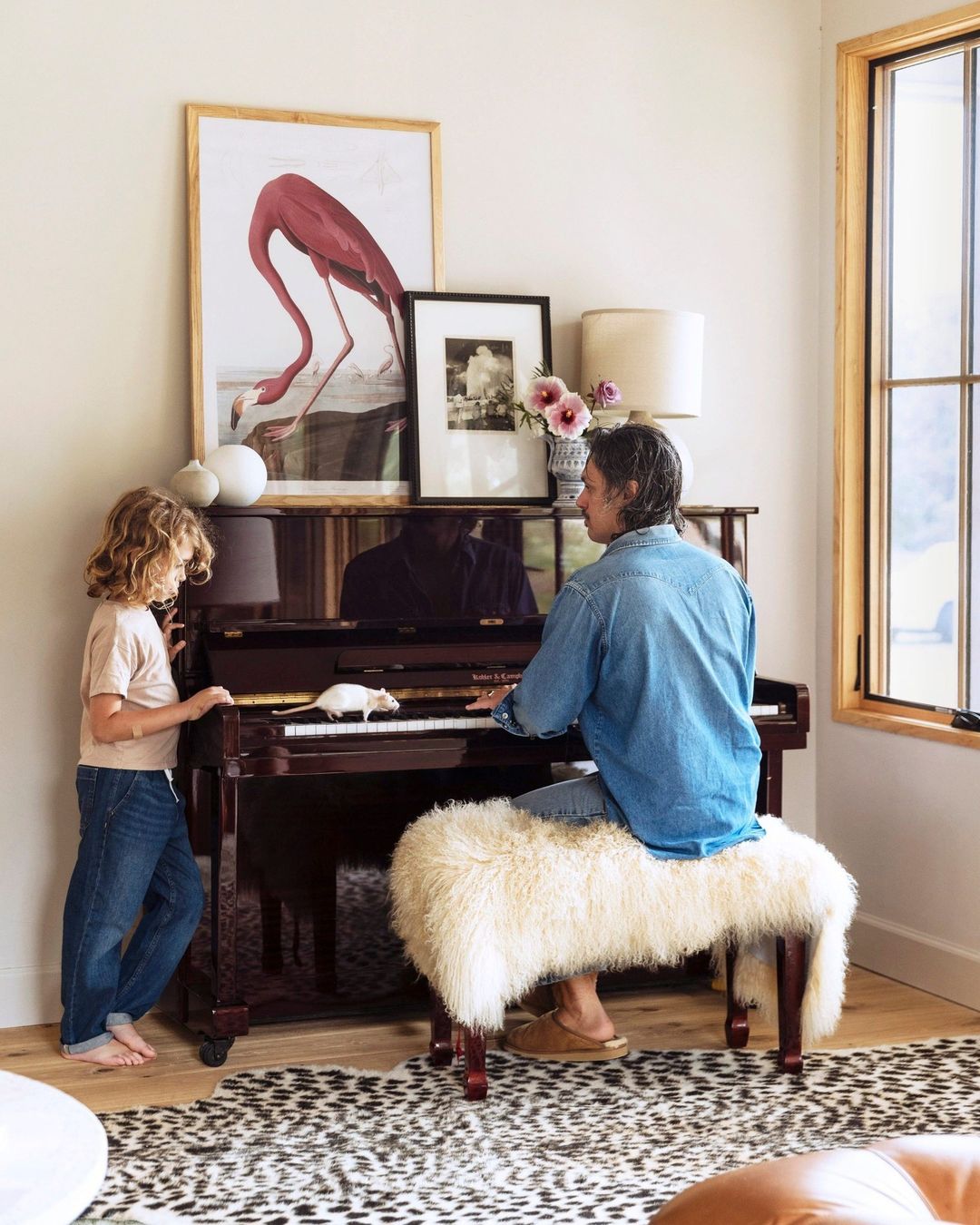 A child and adult playing a piano in a living room with natural light, featuring framed artwork and a furry bench.