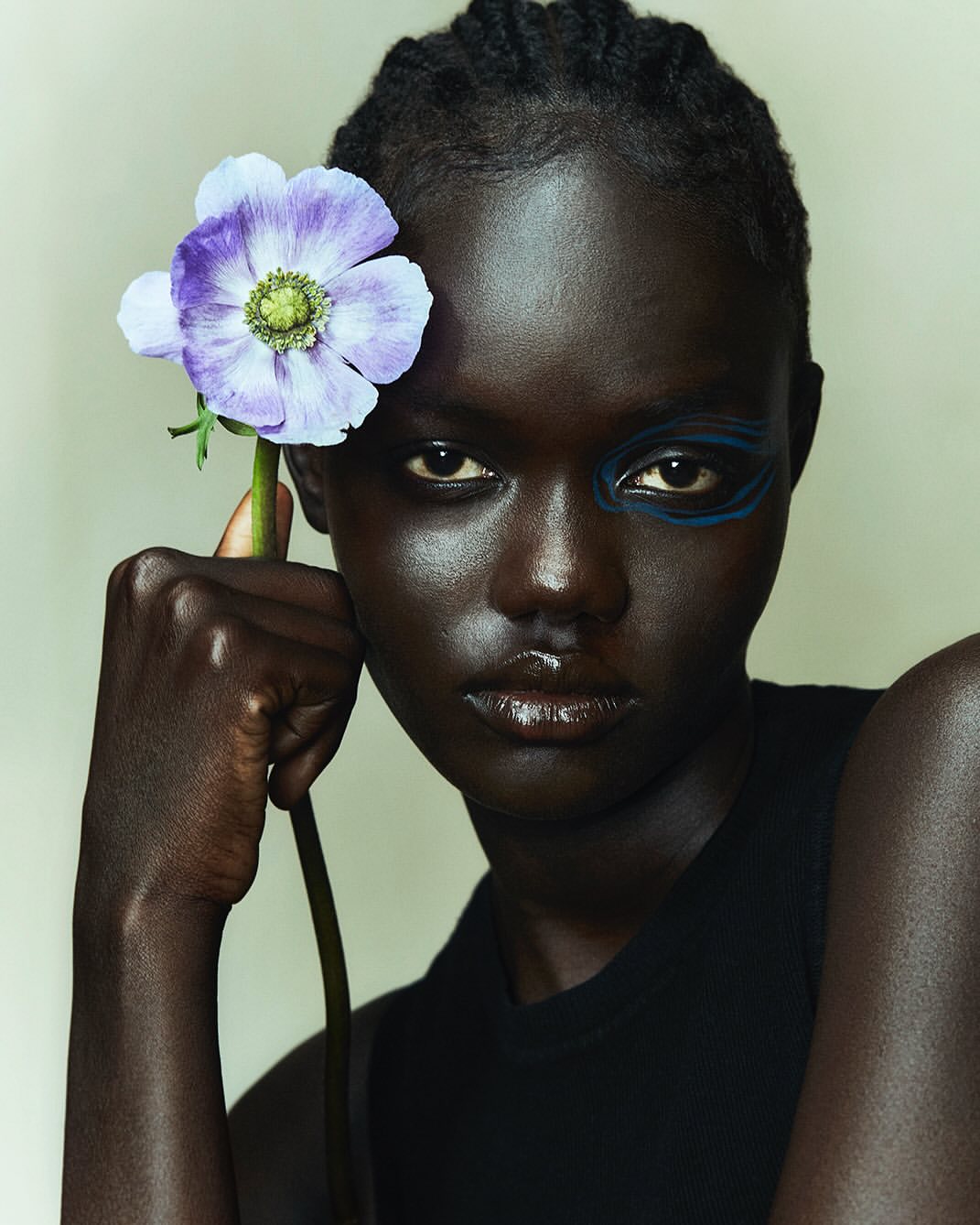 Portrait of a dark-skinned adult female with an anemone flower next to her face and blue makeup on her eye, in a studio environment.