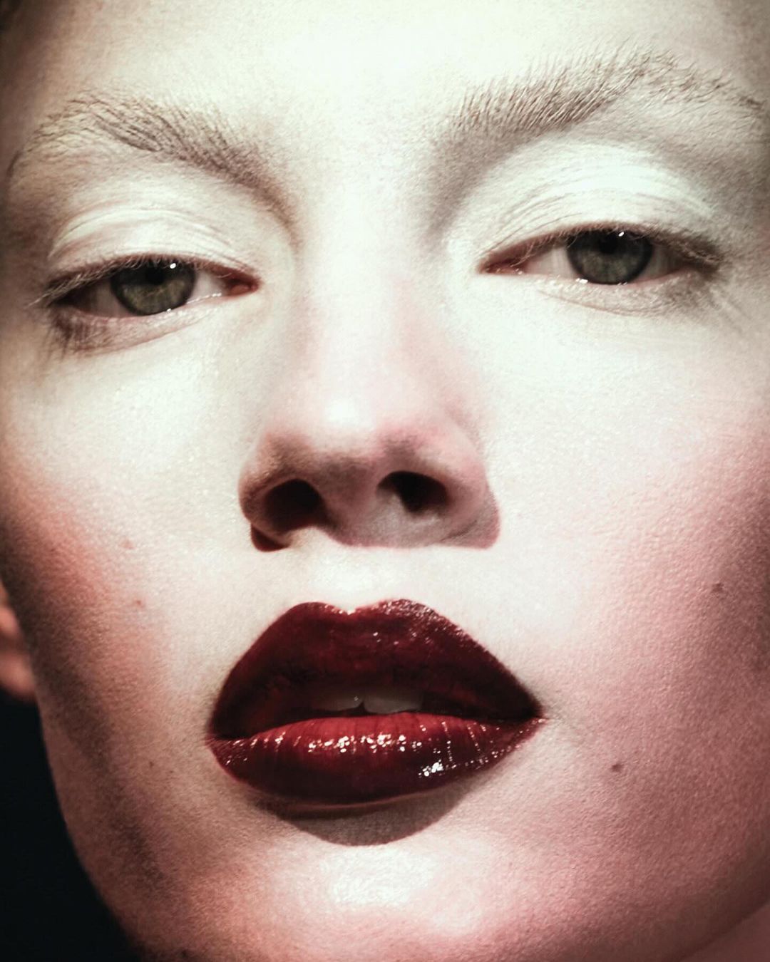 Close-up beauty shot of a fair-skinned person with bleached eyebrows, glossy dark red lips, and fair skin in a dark setting.