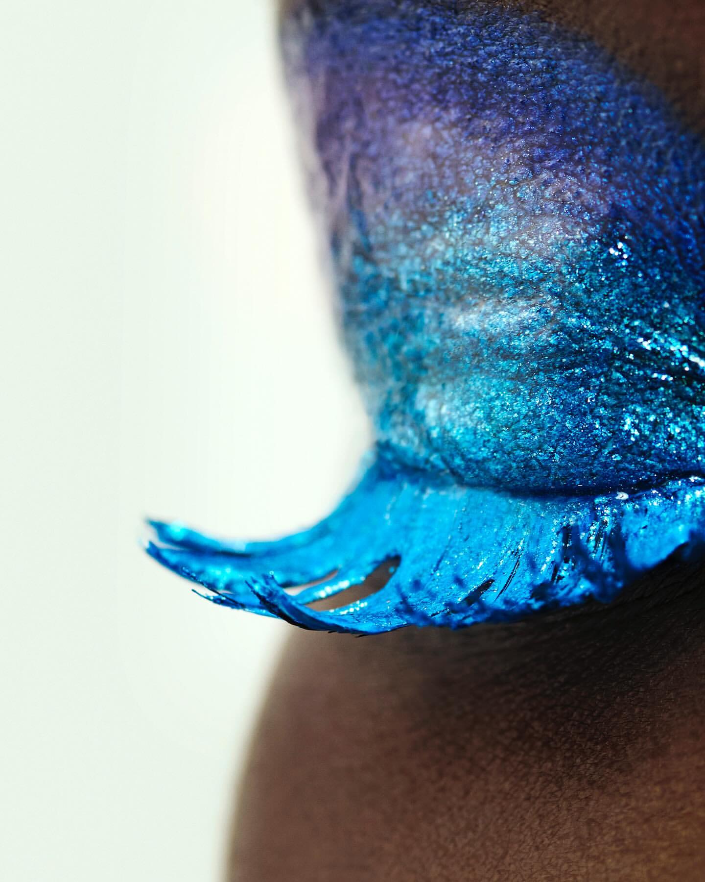 Close-up shot of a model with dark skin and vibrant blue and purple face paint, creating a shimmering and artistic makeup effect.