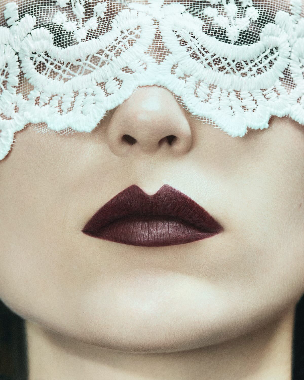 Close-up portrait of a woman's face with dark lipstick and a white lace veil partially covering her eyes and forehead.