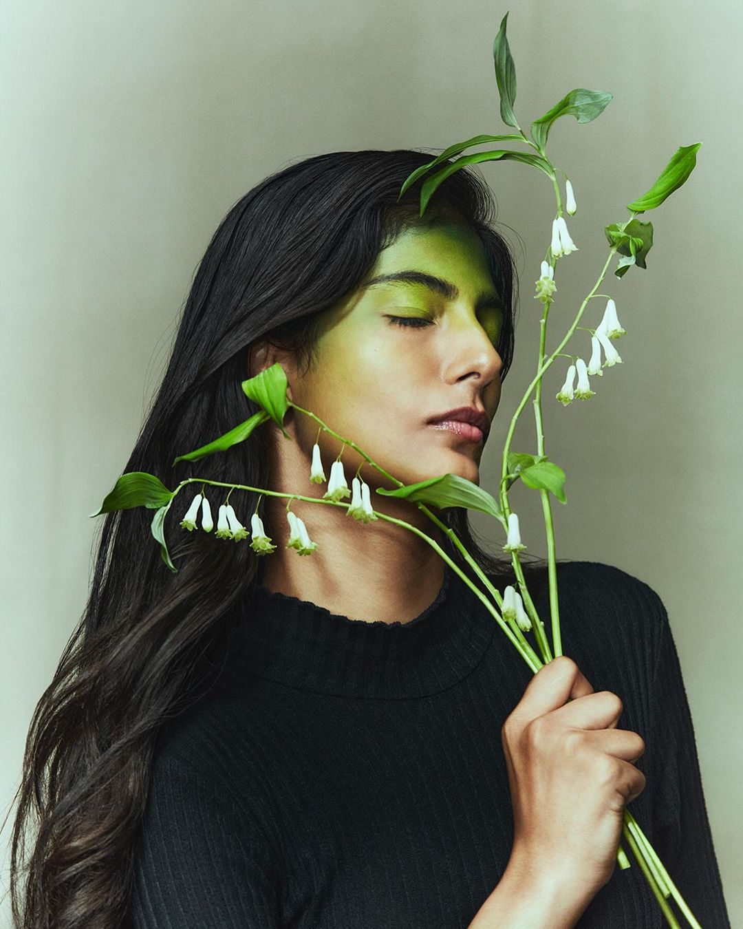 Portrait of a woman with green face paint holding white flowers, wearing a black sweater, set against a neutral background.