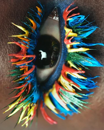 Close-up studio shot of an eye featuring colorful paint on eyelashes and surrounding skin, dark eye, and artistic makeup.