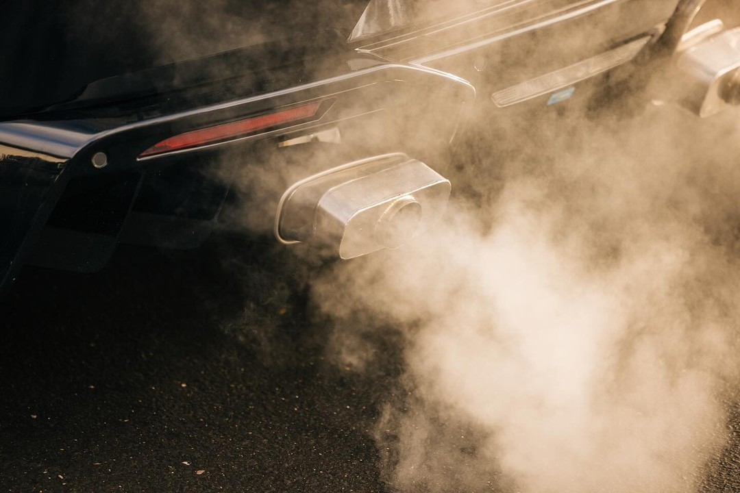Rear view of a black car emitting exhaust fumes from a chrome exhaust pipe. Smoke fills the frame against asphalt.