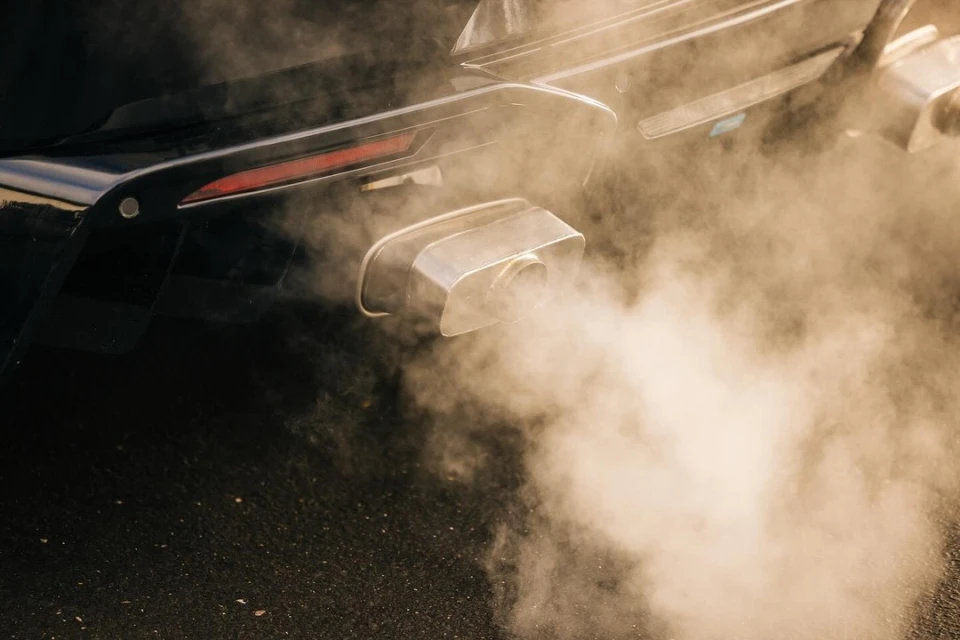 Rear view of a black car emitting exhaust fumes from a chrome exhaust pipe. Smoke fills the frame against asphalt.