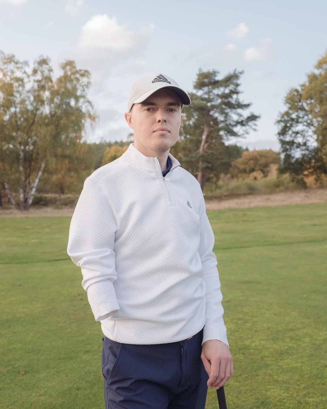 Portrait of a golfer in a white sweater and hat on a green. Trees are in the background under a partly cloudy sky.
