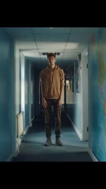 Young man in brown hoodie and patterned pants stands centered in a blue hallway, facing forward with neutral expression, full shot.