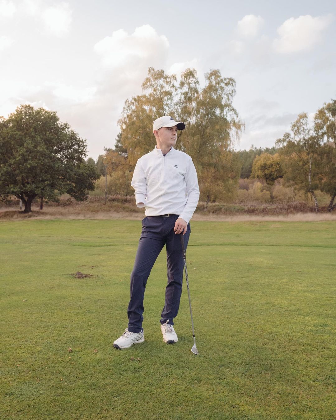 Man with one arm standing on a green golf course holding a golf club and wearing a white long sleeve shirt and a baseball cap.