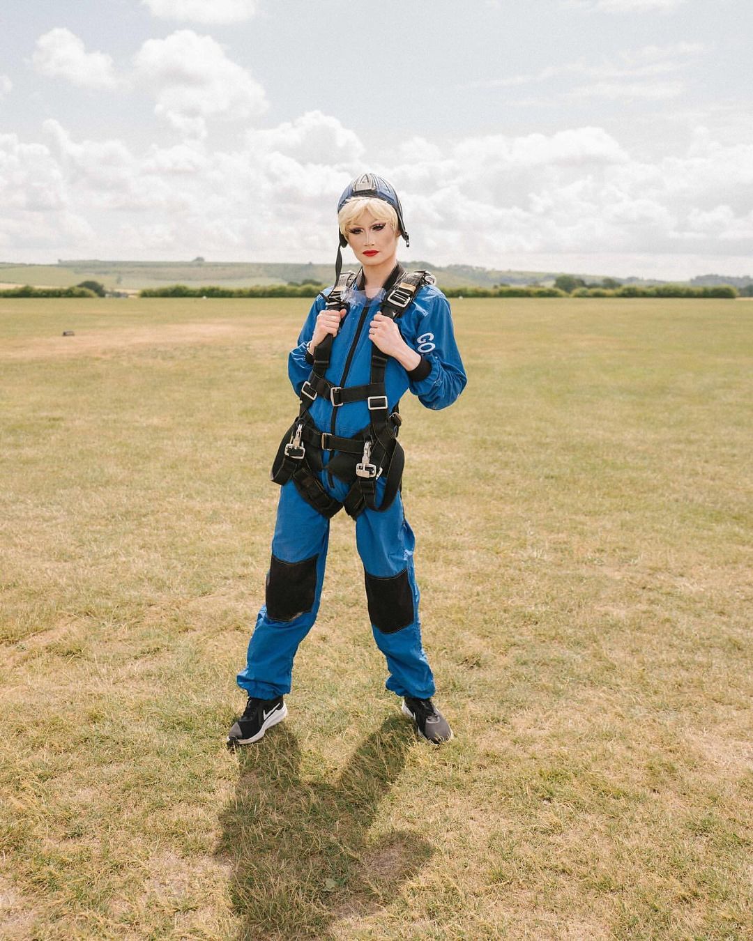 Full body portrait of a blonde adult wearing a blue jumpsuit with a pilot's cap, standing in a grassy field on a sunny day.