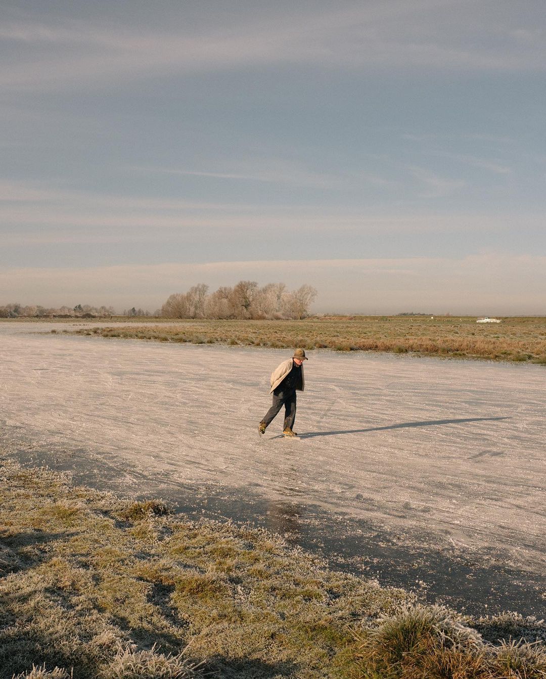 Adult ice skater glides across a frozen river on a winter day with frosty trees and fields along the shoreline under a hazy blue sky.